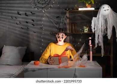 Startup Small Business Entrepreneur Or Freelance Girl In Halloween Makeup Taking Order Online Video Call Using Smart Phone. Young Woman Unpacking Box With Bow In Dark Room. Copy Space