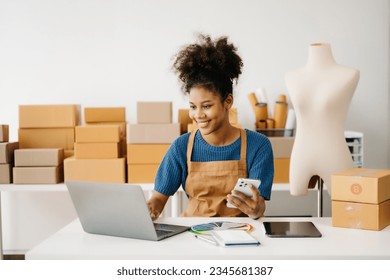 Startup small business SME, Entrepreneur owner African woman using smartphone or tablet taking receive and checking online purchase shopping order to preparing pack product box. 