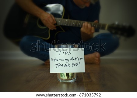 Starving artist guitarist street performer sitting on the sidewalk playing his guitar with a tip cup jar with money in it in front of him in crisp focus.