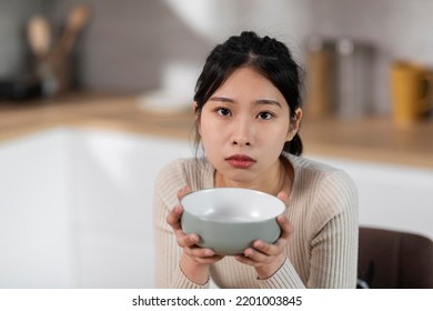 Starving Millennial Asian Woman In Casual Outfit Sitting At Kitchen Table At Home, Holding Empty Plate Next To Her Mouth, Upset Poor Hungry Chinese Lady Craving, Have Nothing To Eat, Copy Space