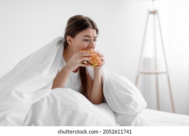 Starving upset hungry cute caucasian millennial woman sitting in bed at home and eating burger suffering from stress, depression, crisis and gluttony. Junk food, substitution, diet and too much meal - Shutterstock ID 2052611741
