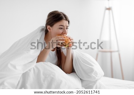 Starving upset hungry cute caucasian millennial woman sitting in bed at home and eating burger suffering from stress, depression, crisis and gluttony. Junk food, substitution, diet and too much meal