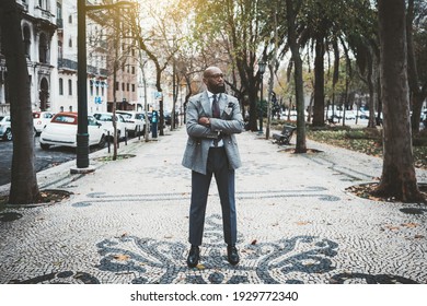 A stately mature hairless barbate black man entrepreneur in a dapper custom made suit is standing with hands crossed on the paving stone in the middle of a city alleyway surrounded by trees and cars