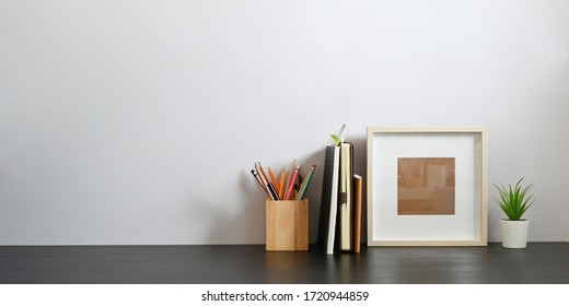 Stationary In Wooden Pencil Holder Putting On Wooden Working Desk That Surrounded By Books, Notebook, Empty Picture Frame And Potted Plant Over Sitting Room White Wall As Background.