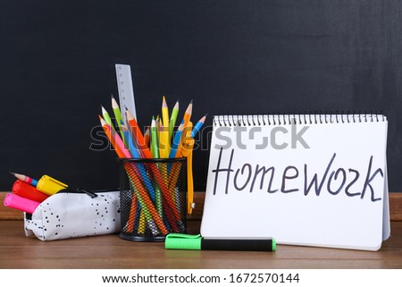 Stationery and notebook with inscription HOMEWORK on wooden table near blackboard
