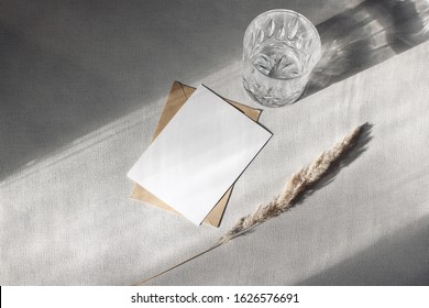 Stationery still life scene. Glass of water, dry grass and craft envelope on gray table background in sunlight. Blank paper card, invitation mockup scene with long shadows. Flat lay, top view.