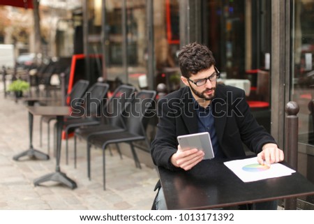 Statistician working with diagram document papers at cafe table outside in  . Hardworking man wears  dark blue shirt. Concept of using modern gadgets and Internet connection for job. 