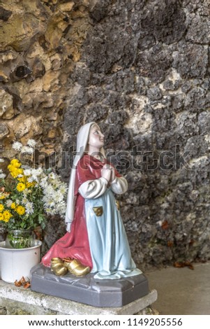Statue of Bernadette praying to Our Lady of Lourdes with flowers next to her and rock wall behind her, wonderful day in south Limburg in the Netherlands