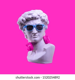 Statue. Earphone on a pink background. Gypsum statue of David's head. Creative. Plaster statue of David's head in blue sunglasses. Minimal concept art.