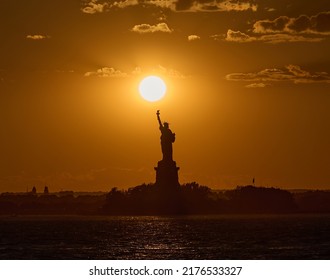 The Statue Of Liberty At Sunset. The Sun Is A Bright Disc Sitting Right Above Liberty's Torch, As Seen From Across New York Harbor.