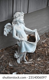Statue of a little angel girl leaning up against a dark grey wall while standing in dirt. The angel is holding up her dress to make something like a basket to carry things in. Very cute. 
