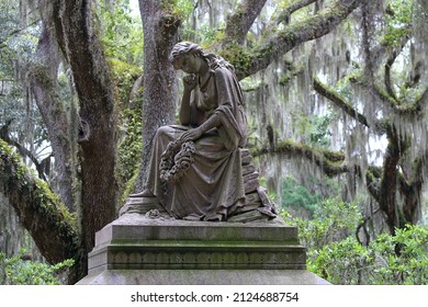 Statue monument of historic woman holding a wreath in a landscape of Spanish moss trees in outdoor cemetery graveyard.