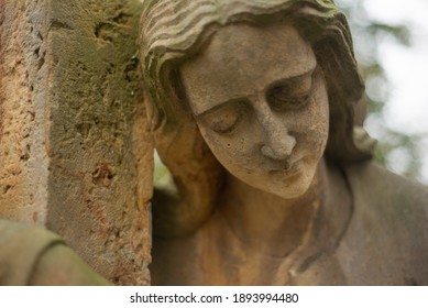 Statue Of A Sad Woman On A Cementery Mourning Over The Grave. Closeup Of Face Of Statue Looking Like A Female Angel. Hand Of Woman Is Hugging A Stone Column Next To Her.