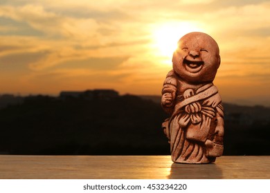 Statue of smiling and laughing Buddha against a sunset background.