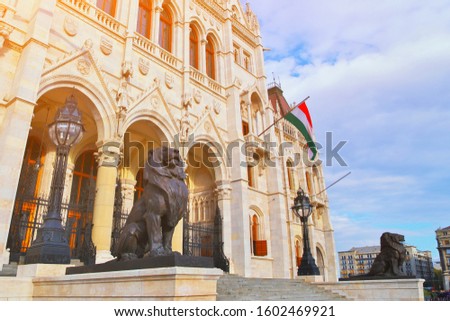 Statues of lion sculpture at the entrance to parliament famous building in Budapest, Hungary. Hungarian flag on blue clouds sky on sunset. Popular travel destination in Budapest, capital of Hungary