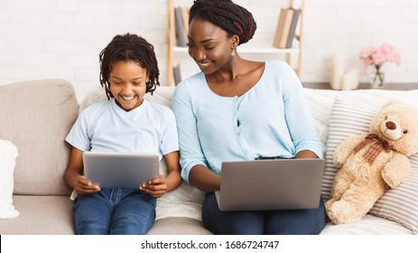 Stay at home african mom working remotely on laptop and helping her child with homework, using tablet and laptop
