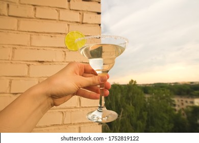 Stay At Home Balcony Cocktail Party. Classic Home-made Margarita Alcohol Drink With Lime And Salt In A Hand, Trees Sky And Residential Area Houses View. Good For Website Or Blog Recipes Or Articles.