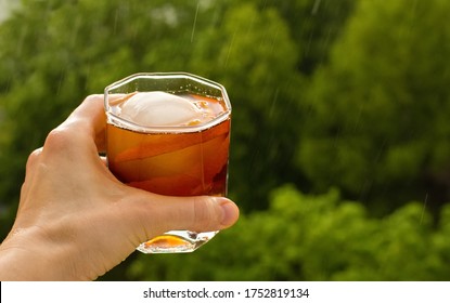 Stay At Home Balcony Cocktail Party. Classic Home-made Old Fashioned Alcohol Drink With Ice Ball And Orange Zest, Trees Sky And Rain Drops. Good For Website Or Blog Recipes Articles. Free Copy Space