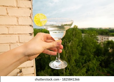 Stay At Home Balcony Cocktail Party. Classic Home-made Margarita Alcohol Drink With Lime And Salt In A Hand, Trees Sky And Residential Area Houses View. Good For Website Or Blog Recipes Or Articles.