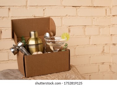 Stay At Home Bar Cocktail Party. Classic Margarita Cocktail And Bartender Stuff Parcel In A Brown Craft Cardboard Box. Gift To Mixology Lover. Beige Brick Wall Background. Tender Retro Hipster Colors.