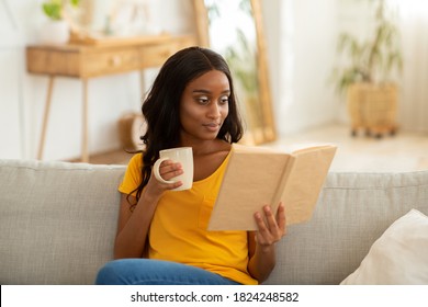 Stay home hobbies concept. Attractive millennial black lady reading book with cup of hot beverage in living room. Focused African American woman with aromatic drink absorbed in intriguing story - Shutterstock ID 1824248582