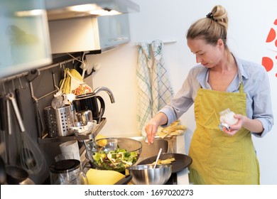 Stay At Home Housewife Woman Cooking In Kitchen, Salting Dish In A Saucepan, Preparing Food For Family Dinner. Blonde Caucasian Model In Her 30is, Real Cooking Not Looking In Camera.