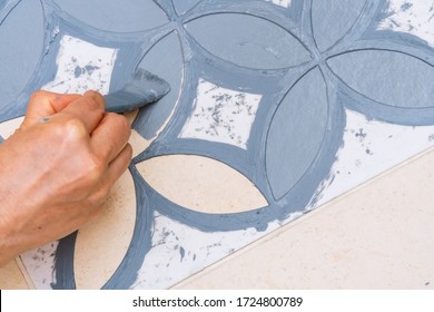 Das Konzept der Heim- und Wohnungsverbesserung: Nahe oben und oben zeigt eine Hand, die eine Bürste hält, eine dekorative Vorlage auf den Bodenfliesen mit einer Vintage-Musterschablone grau