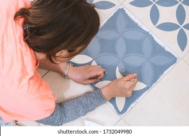 Das Konzept der Heim- und Wohnungsverbesserung: Nahe und obere Ansicht einer Frau, die eine Bürste hält, um eine dekorative Vorlage auf den Bodenfliesen durch Verwendung eines generischen türkischen Musterstollen grau zu malen