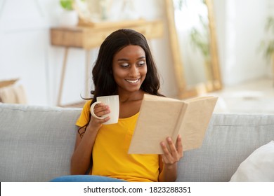 Stay home leisure activities. Lovely young black woman reading book with cup of hot drink in living room. Smiling African American lady with aromatic beverage focused on interesting story - Shutterstock ID 1823288351