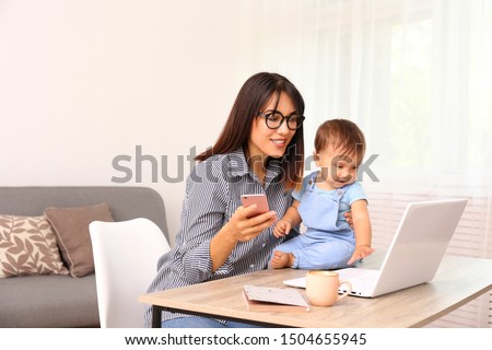 Photo of Stay at home mom working remotely on laptop while taking care of her baby. Young mother on maternity leave trying to freelance by the desk with toddler child. Close up, copy space, background.