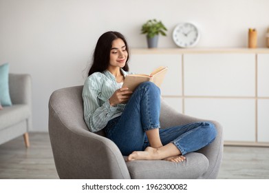 Stay home pastimes. Millennial Indian woman sitting in cozy armchair with open book indoors. Beautiful young lady reading exciting story, enjoying lazy morning, having relaxing weekend - Shutterstock ID 1962300082