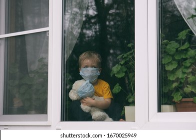 Stay at home quarantine coronavirus pandemic prevention. Sad child and his teddy bear both in protective medical masks sits on windowsill and looks out window. View from street. Prevention epidemic.