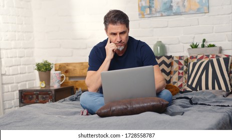 Quédese en casa y trabaje en la oficina del hogar - Hombre informal sentado en la cama trabajando en línea desde casa con computadora portátil.