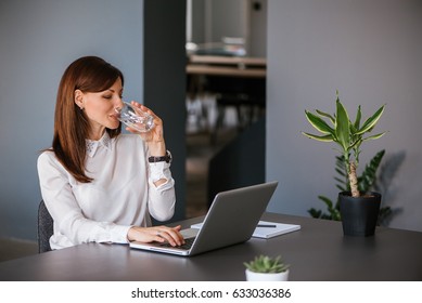 Stay hydrated. Pretty young woman in the office drinking water while working. Drinking from glass.