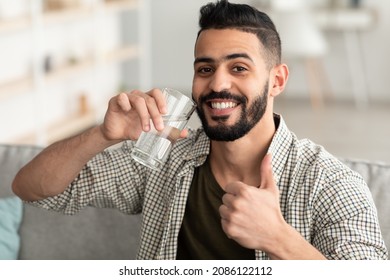Stay hydrated. Smiling Arab guy drinking mineral water from glass and showing thumb up gesture at home. Thirsty young middle Eastern man enjoying refreshing drink in living room