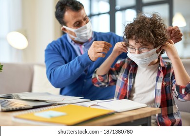 Stay Safe. Middle Aged Hispanic Father Helps His Son, School Boy Wearing Protective Mask While Sitting At The Desk Together, Using Laptop And Having Online Lesson Indoors