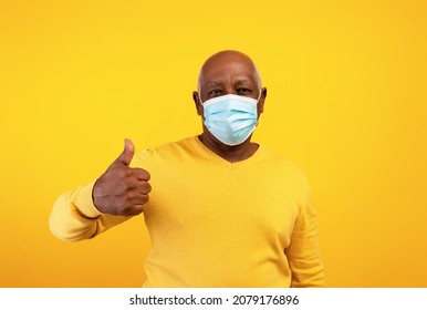 Stay Safe. Senior Black Man In Medical Protective Mask Showing Thumb Up On Orange Studio Background. Elderly African American Male Supporting Anti Covid Preventive Measures. Coronavirus Pandemic