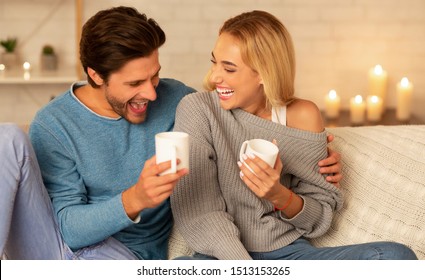 Stay-At-Home Date. Cheerful Couple Laughing Sitting On Couch Drinking Coffee Indoor At Night. Selective Focus