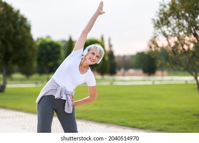 Staying active after retirement. Happy joyful mature retired sportswoman wearing headphones and sportswear doing side stretching exercises with arm over his head, exercising outside in city park - Shutterstock ID 2040514970