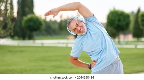 Staying active after retirement. Happy joyful mature retired sportsman wearing headphones and sportswear doing side stretching exercises with arm over his head, exercising outside in city park - Shutterstock ID 2044850414