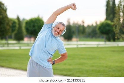 Staying active after retirement. Happy joyful mature retired sportsman wearing headphones and sportswear doing side stretching exercises with arm over his head, exercising outside in city park - Shutterstock ID 2131333251