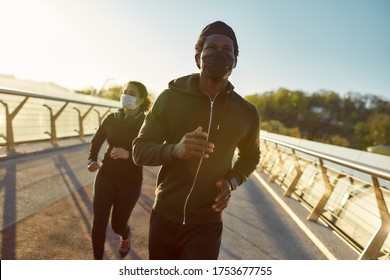 Staying active during quarantine. Young african athletic family in medical masks running on the bridge during a pandemic. Sport and coronavirus. Covid-19. Protection. Social distancing. Couple jogging - Powered by Shutterstock