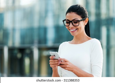 Staying in touch with colleagues. Happy young businesswoman holding mobile phone and smiling while standing outdoors