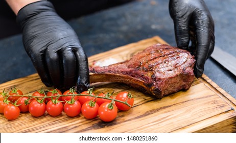 Steakhouse Kitchen. Chef Hands Serving Cowboy Steak With Cherry Tomatoes On Wooden Board.