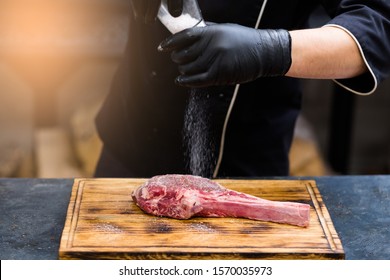 Steakhouse Kitchen. Cropped Shot Of Chef Cooking Cowboy Steak, Salting Raw Beef Bone In Meat On Wooden Board.