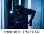 Stealthy criminal wearing black balaclava sneaking into house through window or glass door, using torch at night