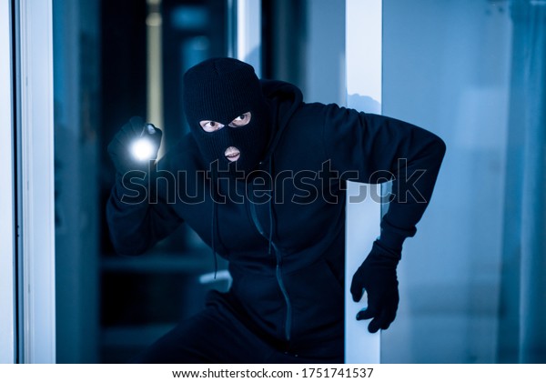 Stealthy criminal\
wearing black balaclava sneaking into house through window or glass\
door, using torch at\
night