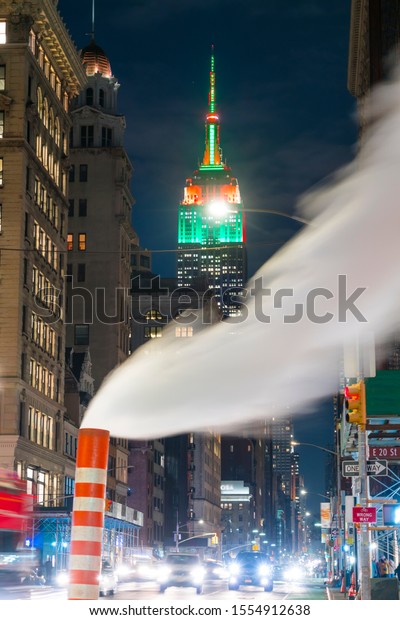 Steam drifts\
among the Fifth Avenue buildings at front of glowing Empire State\
Buildings in Christmas Color at night in New York City NY USA on\
Dec. 26 2018. Cars run on the\
Avenue.