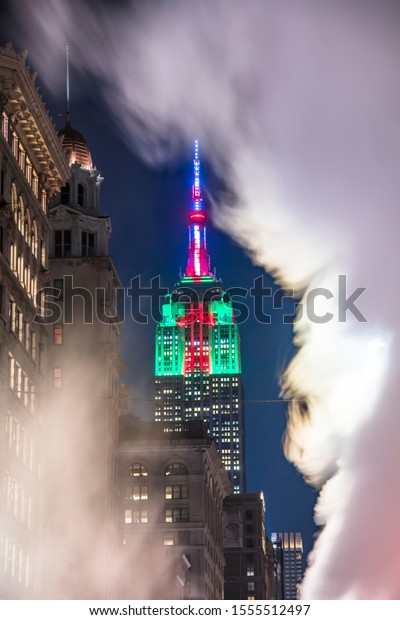 Steam drifts among the Fifth Avenue buildings\
at front of glowing Empire State Buildings in Christmas Color at\
night in New York City NY USA on Dec. 27 2018. People cross and\
cars run on the\
Avenue.\
\
