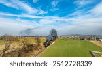 A steam locomotive chugs along railway tracks, billowing dark smoke against a backdrop of lush fields and a clear blue sky. The serene landscape features open fields and distant hills.